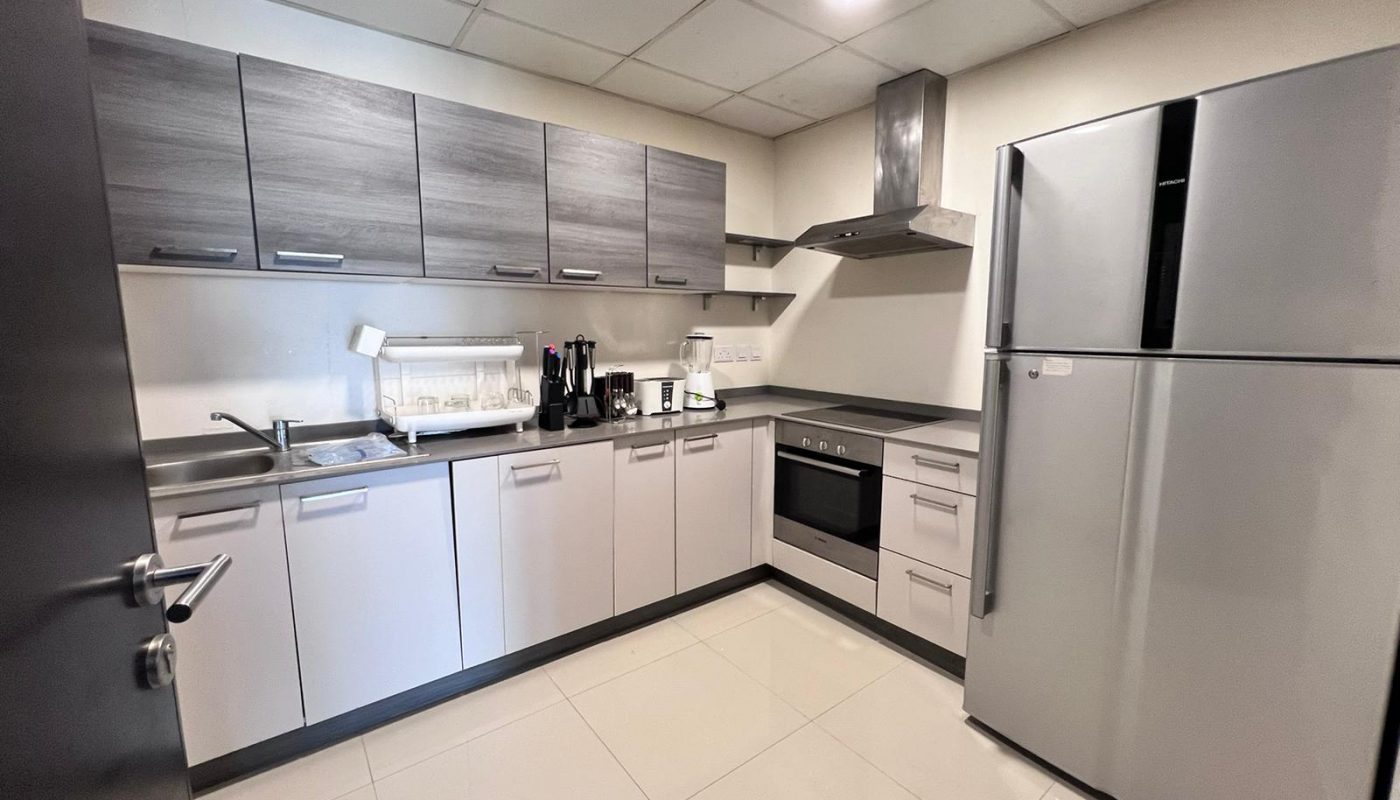 Modern kitchen with light grey cabinets, stainless steel appliances, including a fridge, oven, microwave, and stovetop. The counter has a sink, knife set, toaster, and kettle with white tile flooring. This luxury apartment in Juffair is now on sale!