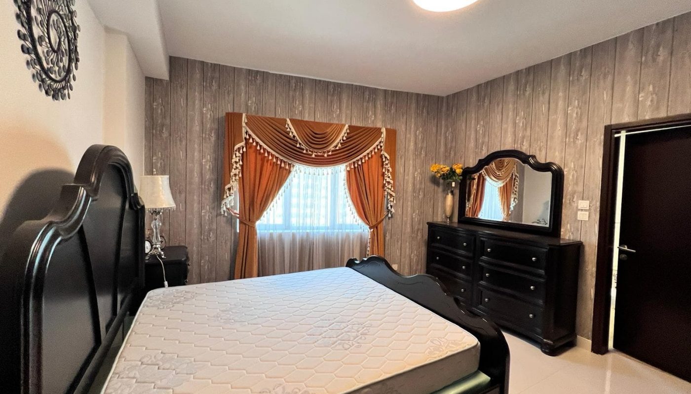 Bedroom in a luxury apartment in Juffair, featuring dark wooden furniture including a bed frame, dresser, and mirror. The bed has a bare mattress. The window is adorned with orange and white drapes with a valance. The walls are covered in textured wallpaper. Available for sale now.