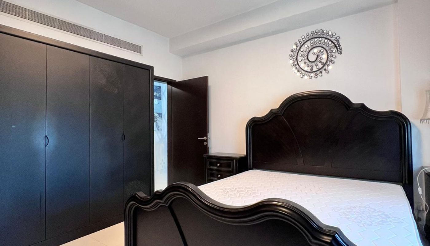 A bedroom with a dark wooden bed, white bedding, black wardrobes, a nightstand, and a round wall decor above the bed. The room has white walls and a closed door.