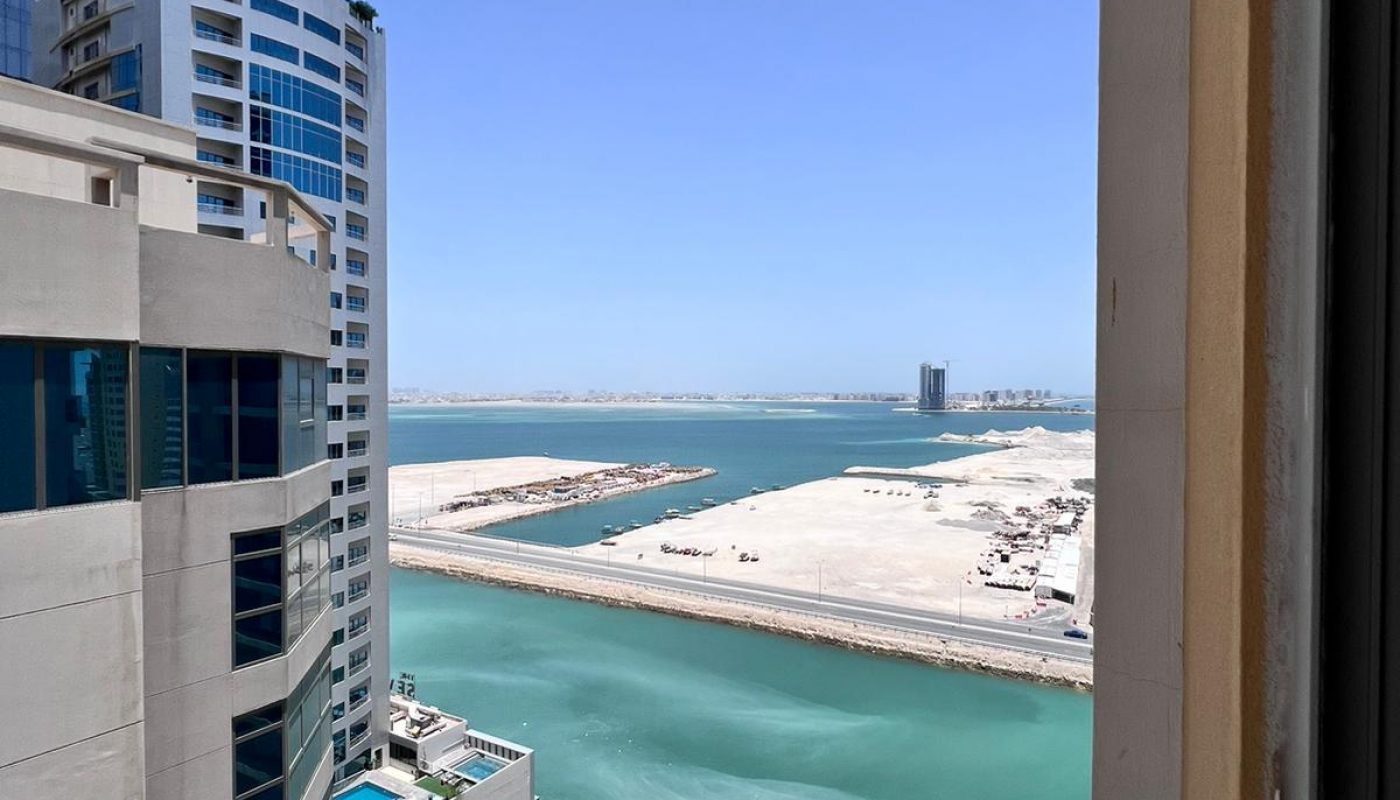 A view from a high-rise window shows turquoise water, sandy land, several modern buildings, and a few parked cars under a clear blue sky.