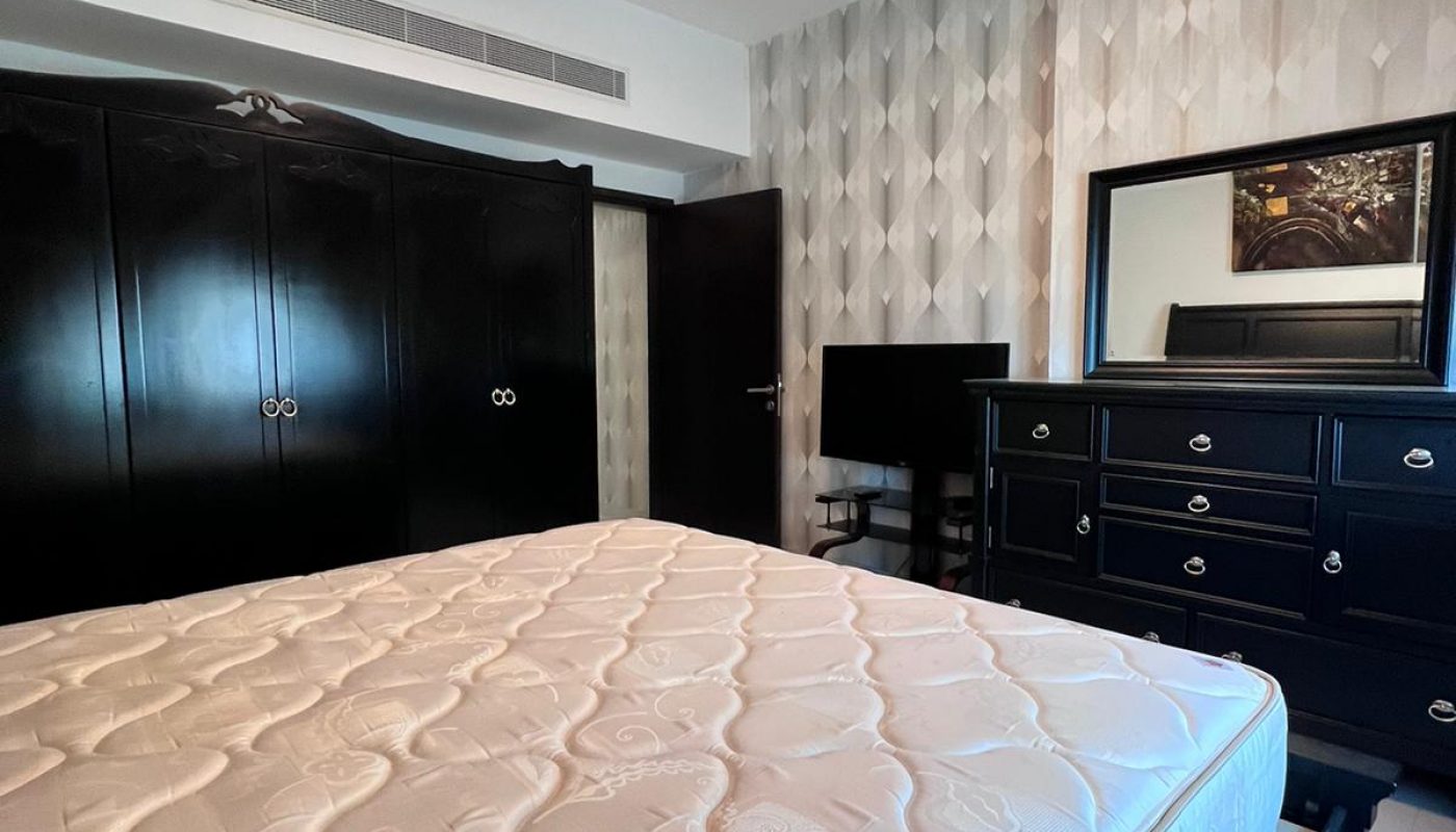 A bedroom with a bare mattress, black wardrobe, black dresser with mirror, a TV on a stand, and patterned wallpaper.
