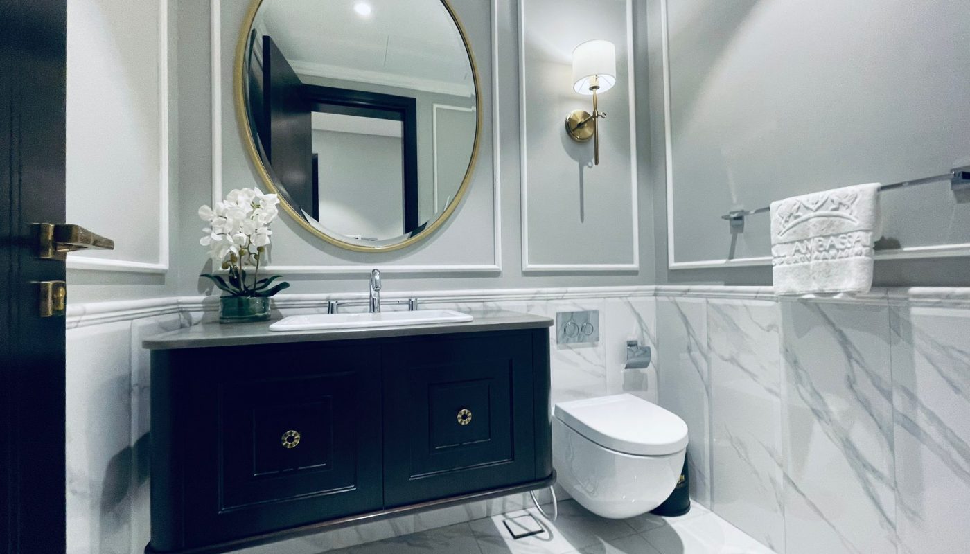 This luxury apartment in Seef features a modern bathroom with a round mirror, wall-mounted sink with storage, white toilet, marble tiles, wall sconce, and a towel rack holding a folded towel with decorative stitching.