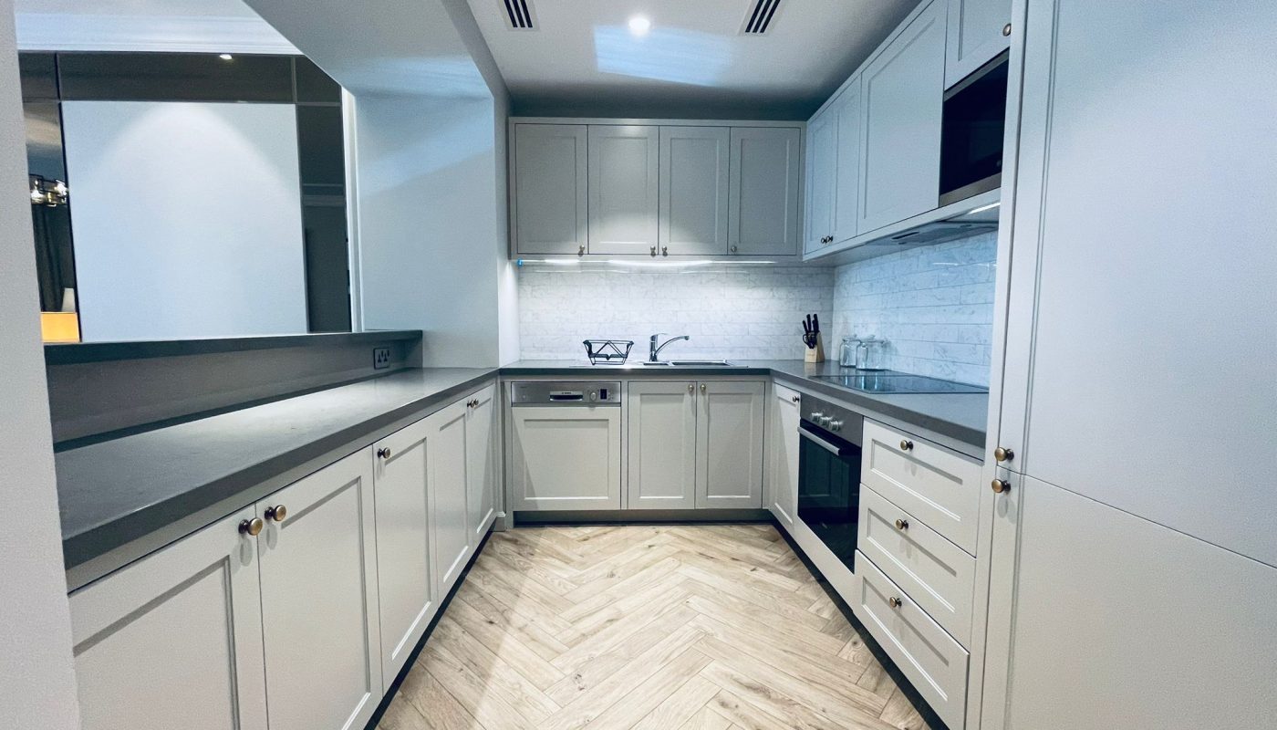 A modern, white kitchen with sleek cabinetry, stainless steel appliances, herringbone-patterned wood flooring, and under-cabinet lighting enhances the luxury of this Seef 2 BR apartment.
