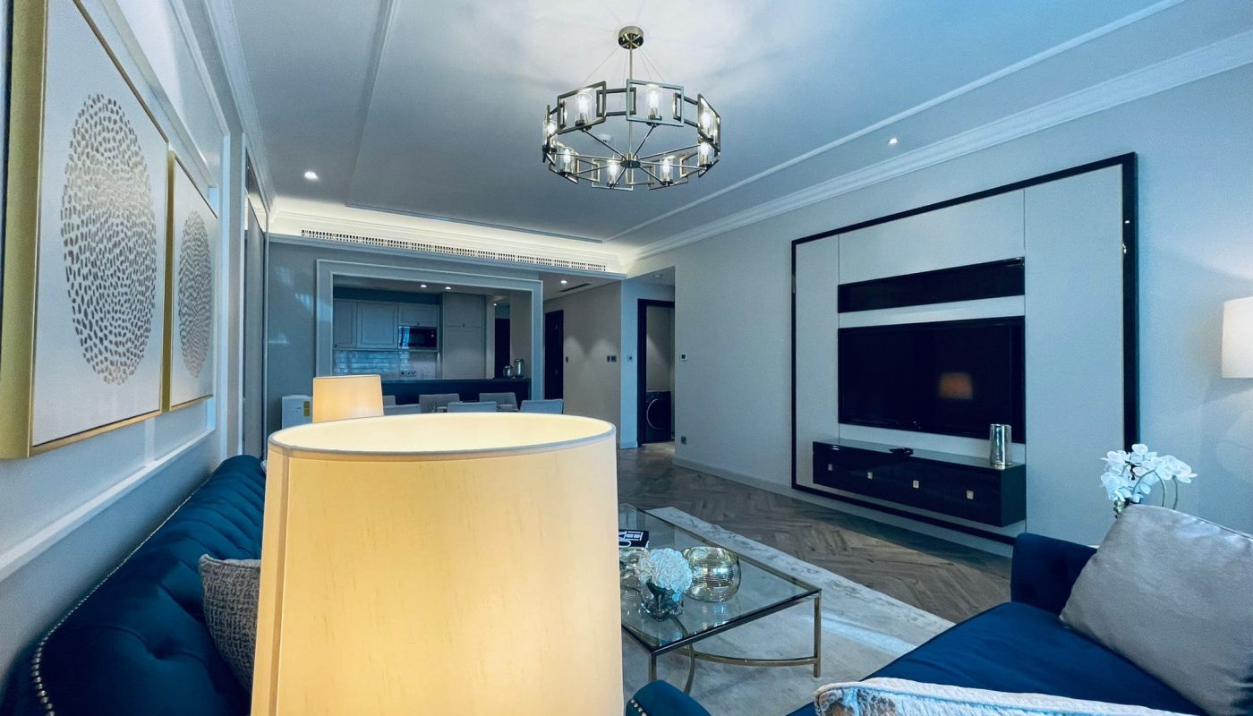 Modern living room in a luxury 2 BR apartment in Seef, featuring blue sofas, a large chandelier, and a wall-mounted TV. The kitchen area is visible in the background. Decor includes framed abstract art and a table with a lamp and flowers.