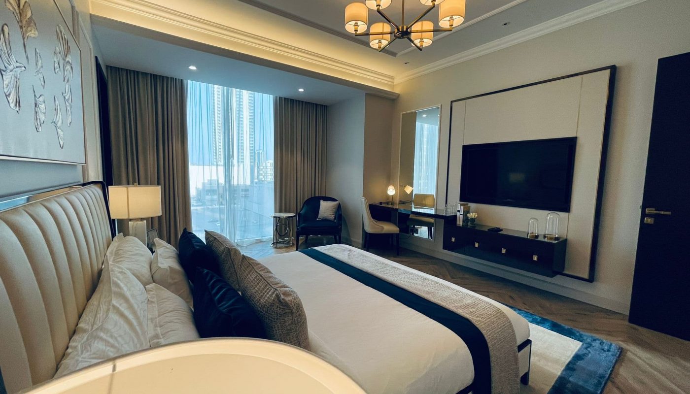 A luxurious 2 BR apartment in Seef, featuring a bed with white linens, a flat-screen TV on the wall, a chandelier, and a city view through large windows with curtains.