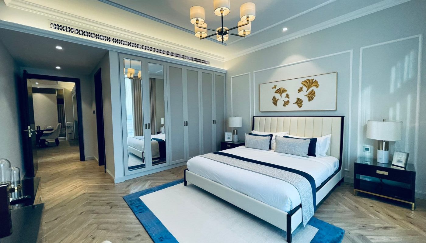 Modern bedroom in a luxury apartment with a large bed, blue accents, mirrored wardrobe doors, bedside tables with lamps, and a chandelier. The room features wooden flooring and a neutral color scheme for an elegant living experience in the heart of Seef.