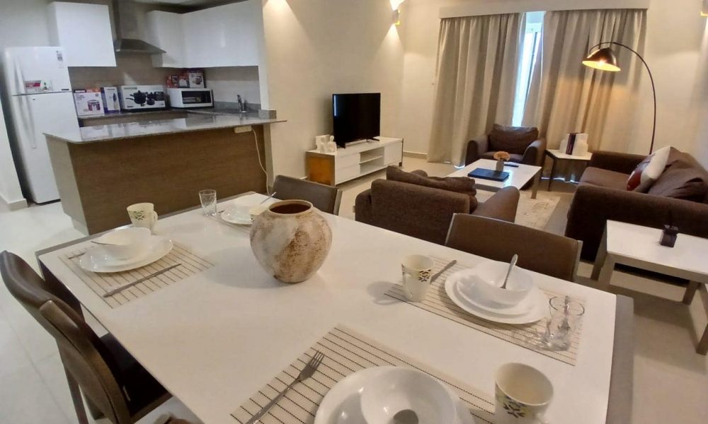 A modern kitchen and living room combination in Amwaj Islands featuring a dining table set for four, a kitchenette with appliances, a comfortable seating area, and contemporary lighting within fully furnished apartments.