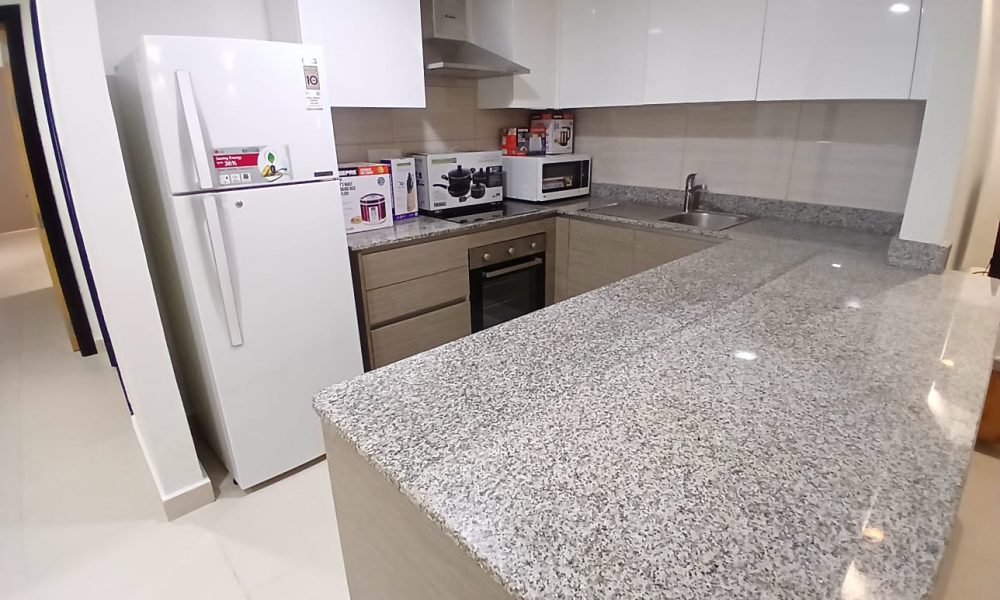 A modern kitchen with white cabinets, a refrigerator, microwave, countertop stove, and granite countertops. The floor is tiled, and the walls are light-colored. This stylish space is ideal for fully furnished apartments in Amwaj Islands.