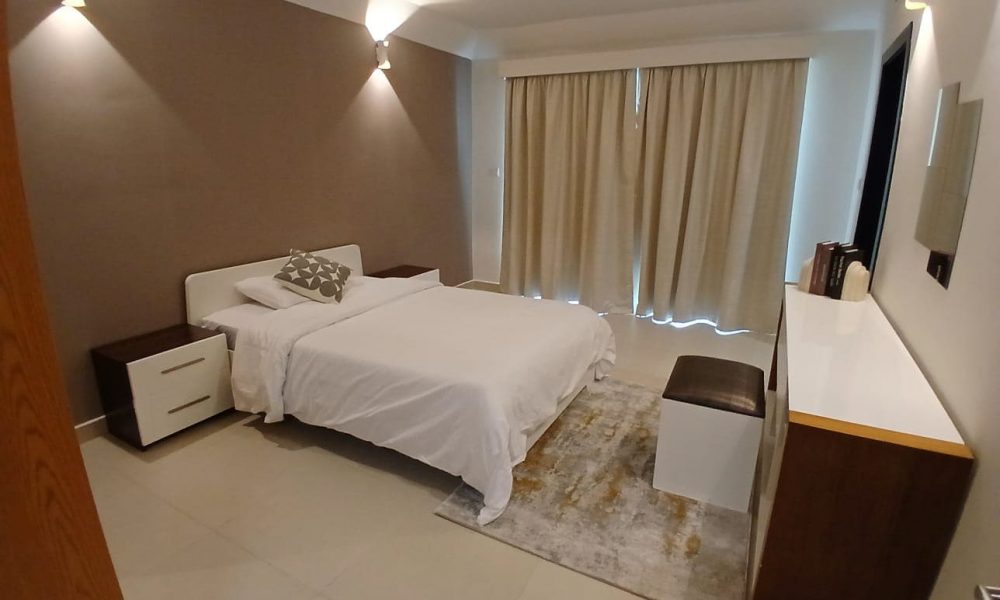 A minimalist bedroom in a fully furnished apartment on Amwaj Islands features a double bed with white linens, two bedside tables, a dresser with a bench, and beige curtains covering a window. The walls are neutral-toned, complemented by modern wall sconces.