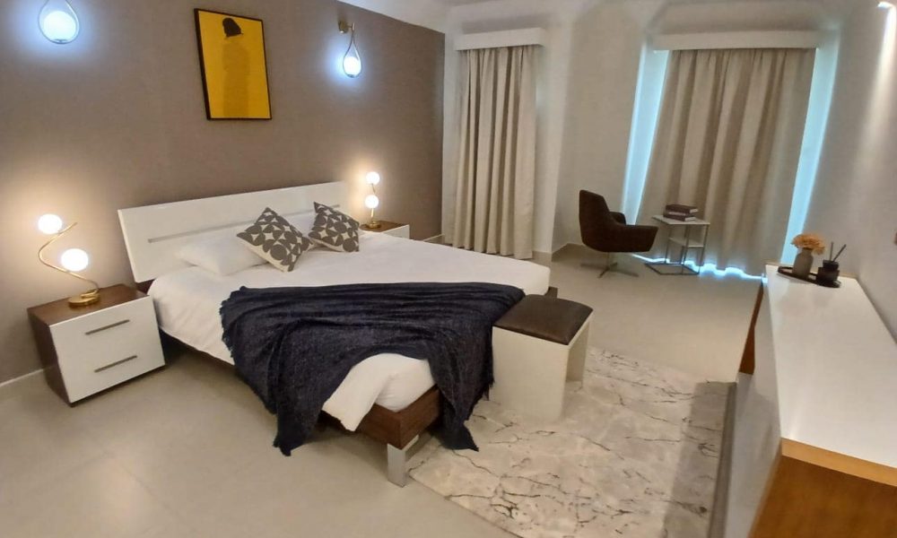 A modern bedroom in Amwaj Islands with a double bed, bedside tables, wall art, and a blue throw. The fully furnished apartment includes a chair and table near the window with curtains drawn. Soft lighting and neutral tones dominate the decor.