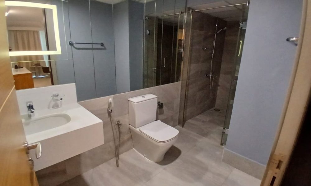 Discover the modern bathroom in our fully furnished apartments at Amwaj Islands, featuring a white sink, toilet, and glass-enclosed shower. The walls are grey, complemented by a large mirror above the sink.