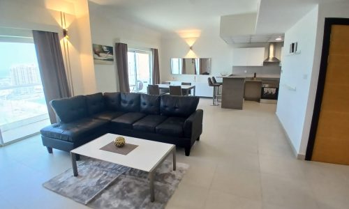 Open-plan living room and kitchen area in this fully furnished flat on Amwaj Islands, featuring a black leather sofa, white coffee table, dining table, and modern kitchen with bar stools. Large windows with curtains allow natural light to fill the space.