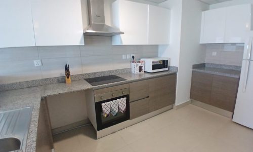 Modern kitchen in a furnished flat on Amwaj Islands features white and wooden cabinets, stainless steel appliances including a stove, microwave, and refrigerator, with a grey countertop and a knife set on the counter.