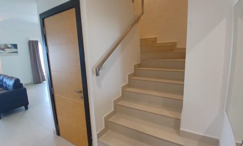 Indoor space with a wooden door on the left, a staircase with a metal handrail leading upwards on the right, and a view of the fully furnished flat's living room with a couch in the background in Amwaj Islands.