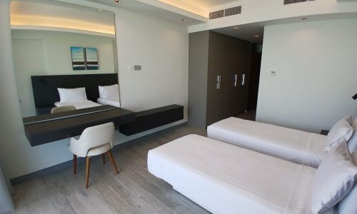 A modern hotel room with two single beds, a wall-mounted desk with a chair, a large mirror, and a closet. The room features minimalist decor with neutral colors and a wooden floor, akin to the stylish apartments you’d find in Diyar Al Muharraq.