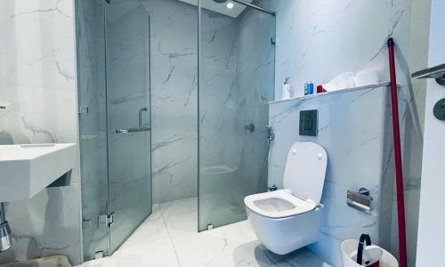 A modern bathroom in an amazing studio for rent in the Seef area, featuring marble walls and flooring, a glass shower enclosure, a white toilet, a wall-mounted sink, and various toiletries on a shelf. The scene also includes a mop and bucket.