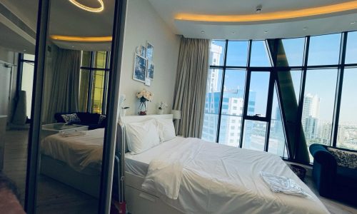 A modern studio for rent in the Seef area features a neatly made bed, large floor-to-ceiling windows, a wall mirror, a small sofa, and stunning cityscape views.