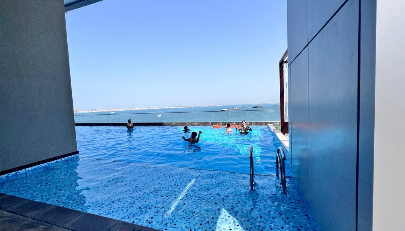 People swim in an infinity pool overlooking the sea on a clear, sunny day, with blue sky and distant boats visible on the horizon.