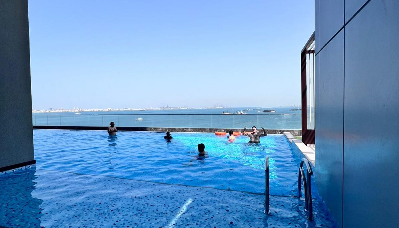 People are swimming in an infinity pool on a building's terrace with a view of the sea and clear blue sky in the background.