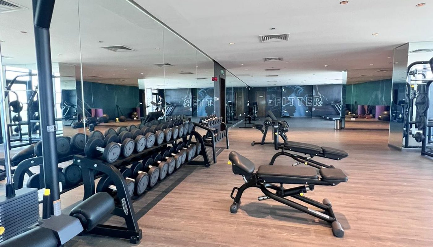 A modern gym with weight racks, benches, mirrors, and exercise equipment on a wooden floor. The room is well-lit with ceiling lights.