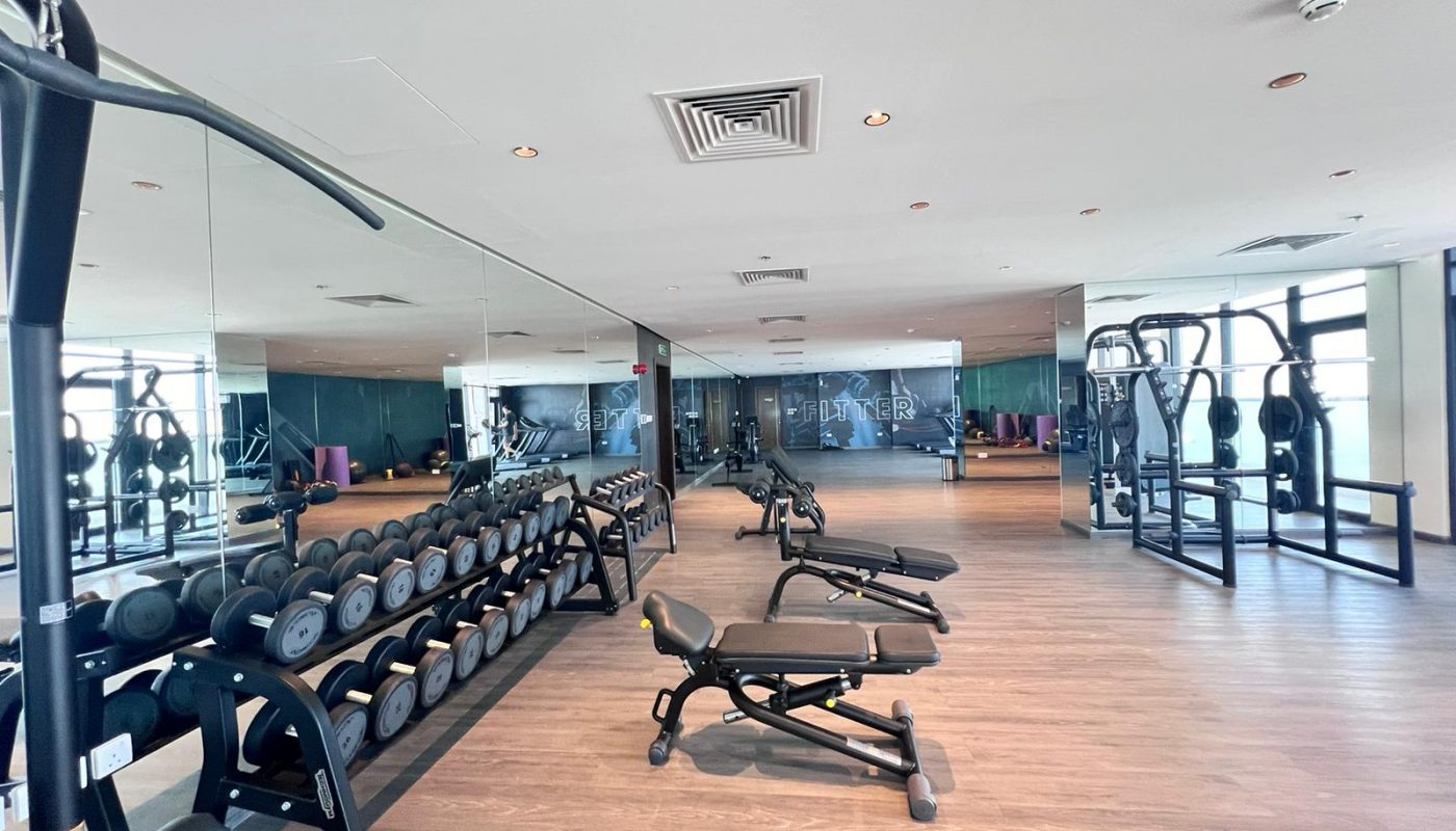 Spacious gym with free weights, benches, exercise machines, mirrors on one wall, and large windows providing natural light.
