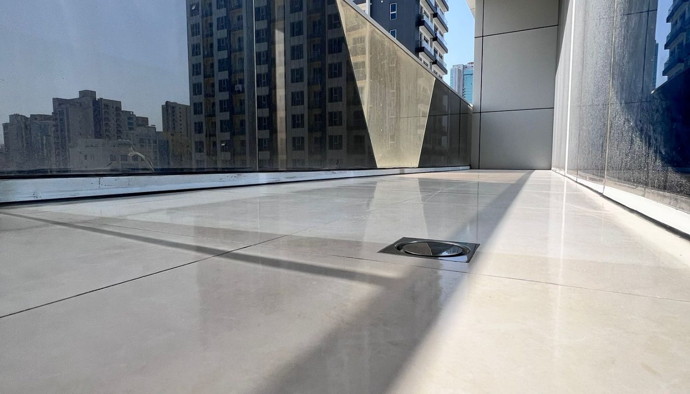 Low-angle view of a modern building’s balcony with glass railing, tiled floor, and city buildings visible in the background under a clear sky.