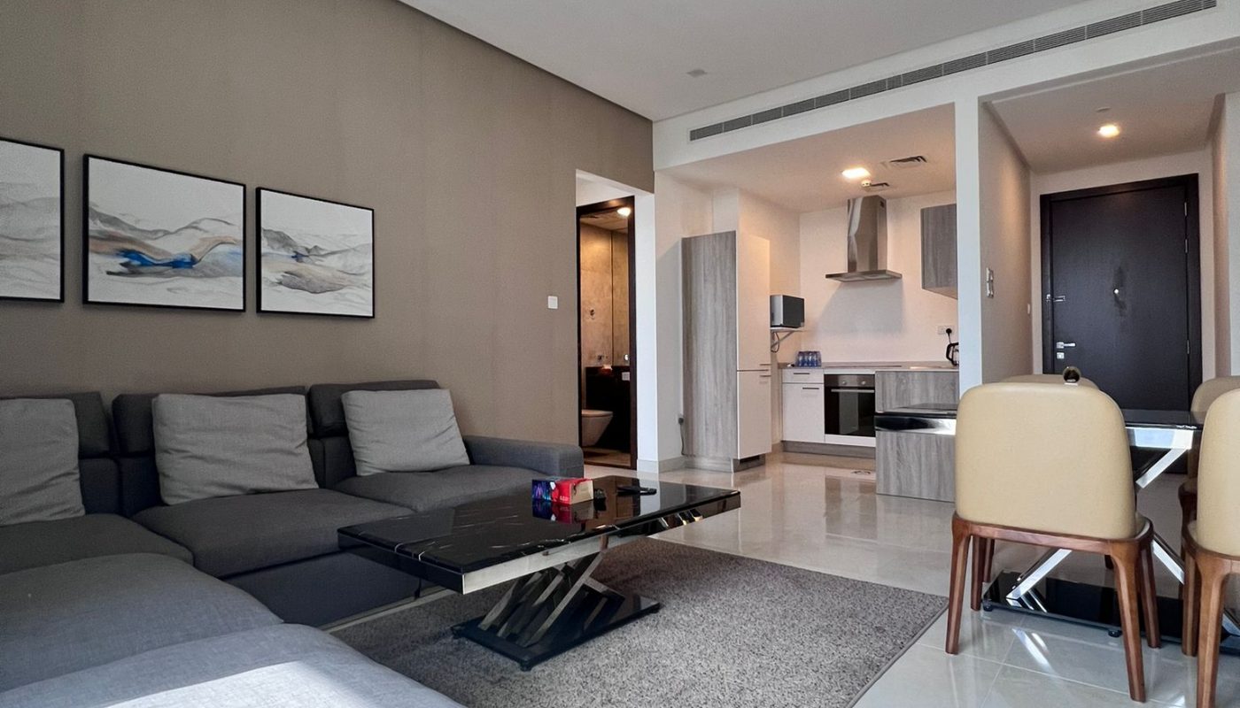 Modern living room with gray sectional sofa, coffee table, wall art, dining table with chairs, and view into kitchen area; sunlight streams in through unseen windows.
