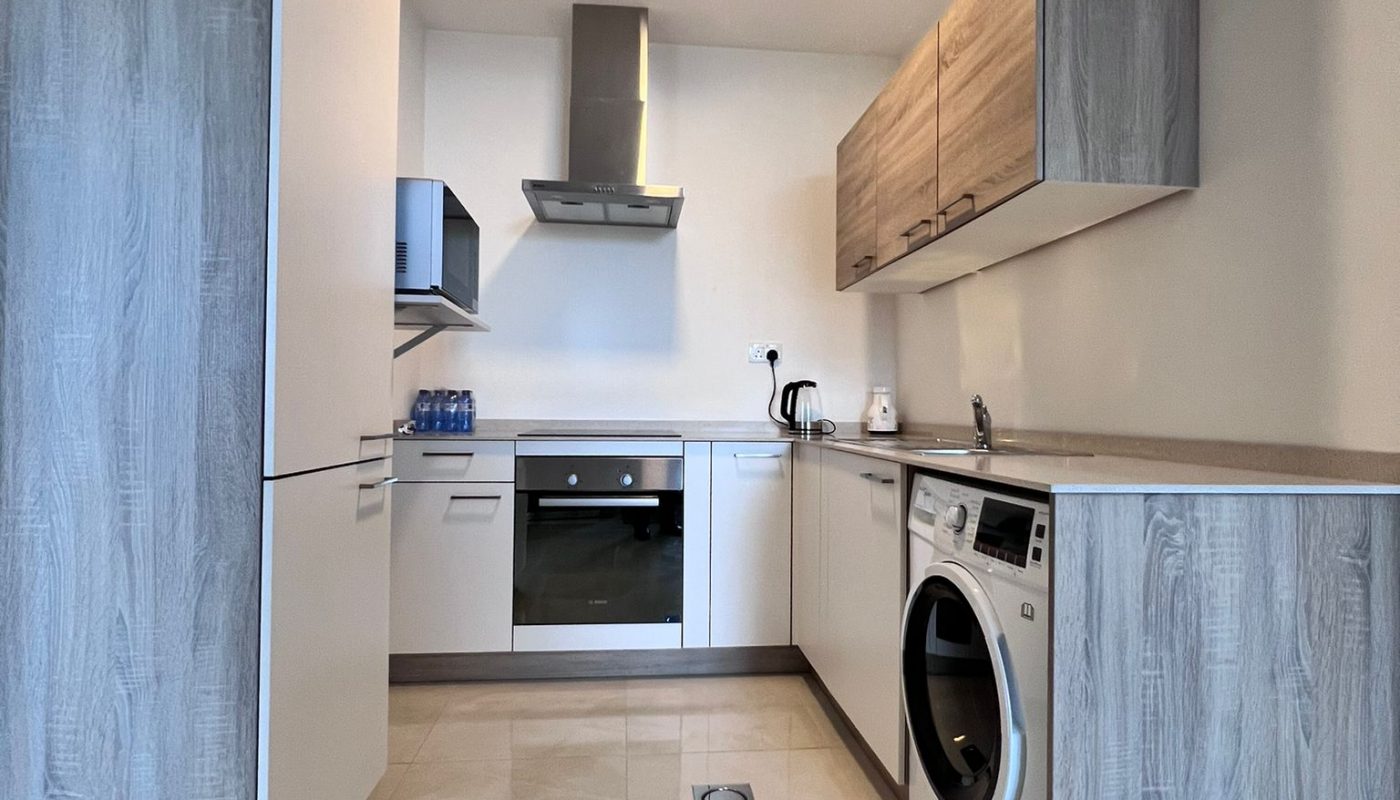 Modern kitchen with wood-finish cabinets, built-in oven, stovetop, washing machine, microwave, kettle, and toaster. Beige tiled floor and neutral walls create a clean, minimalist look.