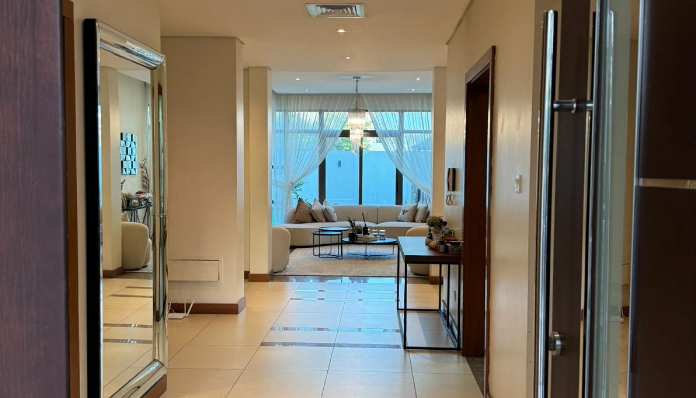View from an open doorway into a bright hallway with tiled floors, a large wall mirror, and a living room with sofas and a coffee table at the far end.