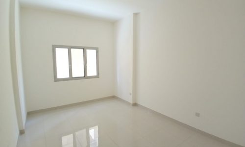 An empty room with white walls, a tiled floor, and a window on one side. The space is well-lit with natural light.