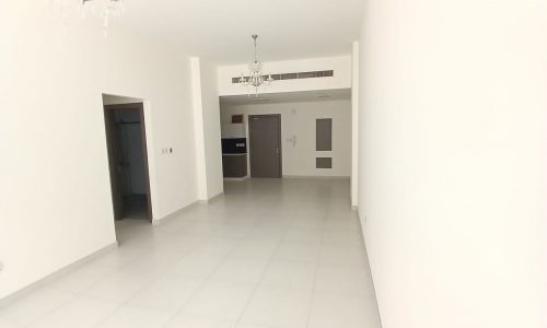 A spacious, empty room with white walls and tiled floor. The room features a chandelier, a door, and hallway entrances on the back wall.