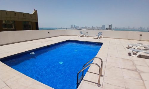 A rooftop pool with blue water surrounded by beige tiles. Four lounge chairs are placed near the pool, and a distant city skyline is visible under a clear sky.