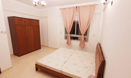 A cozy apartment in Busaiteen featuring a bedroom with a double bed, pink curtains, wooden wardrobe, and beige tiled floor under warm lighting. The best location for comfort and charm.