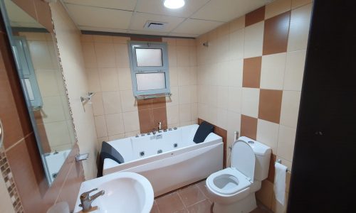 A very good condition bathroom in a Busaiteen apartment features a bathtub, toilet, sink, and mirror. The walls are tiled in shades of beige and brown, with a window above the bathtub allowing natural light to fill the space. This apartment is situated in the best location.