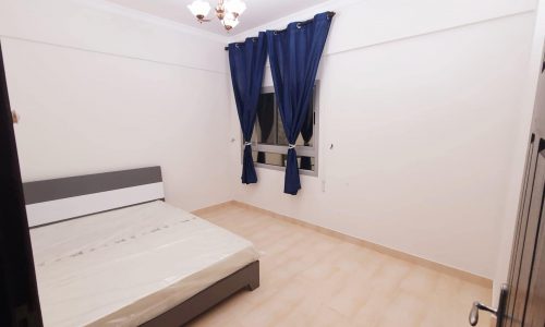 A simple, unfurnished bedroom in a Very Good condition Apartment with a bed frame and mattress, blue curtains covering the window, white walls, beige tiled floor, and a ceiling light fixture. Ideally located in Busaiteen for convenience.