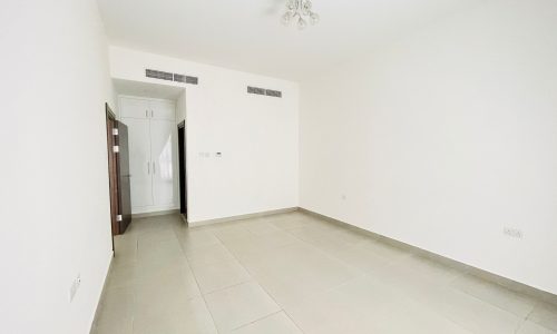 A clean, semi-furnished apartment in Adliya featuring an empty room with white walls, light beige tiled floor, a built-in wardrobe, and a small ceiling light fixture.