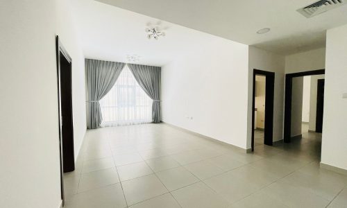 Spacious semi-furnished living room in an Adliya apartment, featuring white walls, tiled floor, a large window with gray curtains, and several doorways leading to other rooms.