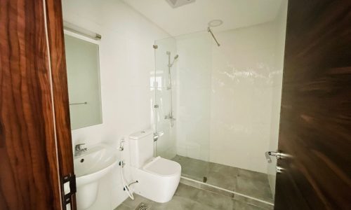 A semi-furnished apartment in Adliya features a modern bathroom with a sink, toilet, and glass-enclosed shower area. The room boasts grey tile flooring, white walls, and a mirror mounted above the sink.