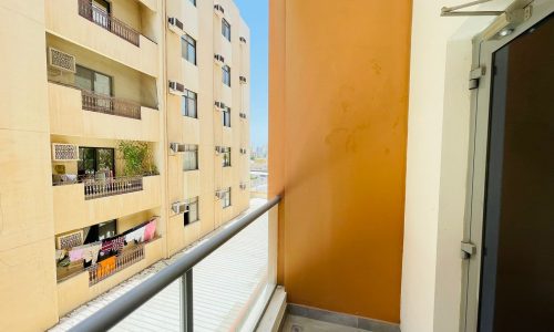 A semi-furnished apartment in Adliya features a small balcony with an orange accent wall, overlooking adjacent apartment buildings. The balcony has a glass railing and is partially visible from the interior through a glass door.