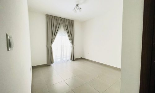 An empty room in a semi-furnished apartment features a tiled floor, beige curtains covering a large window, and a ceiling light fixture, perfect for customizing to your taste in the heart of Adliya.