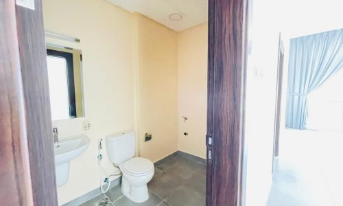 A small bathroom in a semi-furnished apartment in Adliya features a sink and toilet. The room has light beige walls, a square mirror, and a small window. An adjacent room with gray floor tiles and curtains is partially visible.