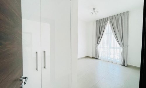 A minimalistic, semi-furnished apartment in Adliya with a tiled floor features large windows covered by light gray curtains. The room includes a white closet with double doors on the left and a light fixture hanging from the ceiling.