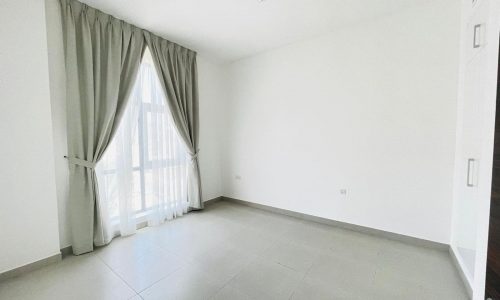 An empty room in a semi-furnished apartment in Adliya, featuring light-colored tiled flooring, white walls, a window with curtains, and a ceiling light fixture.