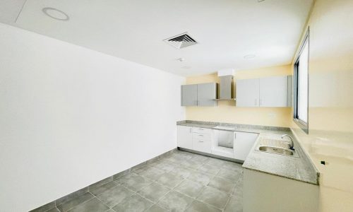 A modern kitchen in a semi-furnished apartment with white upper and lower cabinets, a gray countertop, a stainless steel sink, and a tile floor. The room is brightly lit with natural light from a window on the right, offering a perfect view of Adliya.