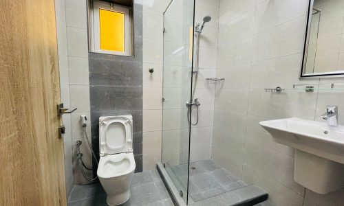 Modern luxury bathroom in Umm AL-Hassam with a toilet, glass-enclosed shower, sink, large mirror, and small window. Grey tiled floor and walls complement the wood door on the left side. Perfectly suited for a 2BR residence.