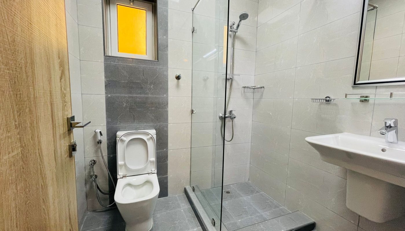 Modern luxury bathroom in Umm AL-Hassam with a toilet, glass-enclosed shower, sink, large mirror, and small window. Grey tiled floor and walls complement the wood door on the left side. Perfectly suited for a 2BR residence.