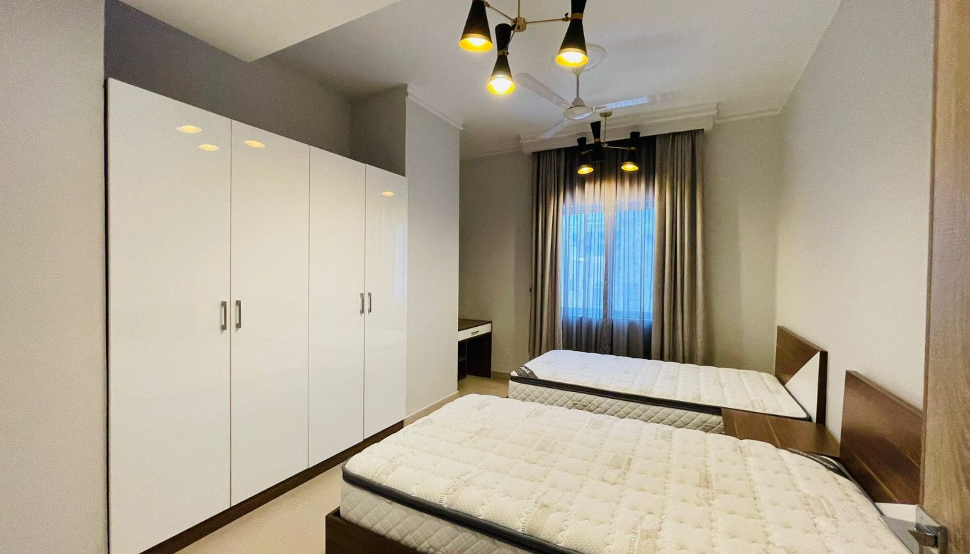 Experience luxury in this 2BR bedroom featuring two beds, a large white wardrobe, beige curtains adorning the window, and a modern ceiling light fixture. Located in Umm AL-Hassam, it's the ultimate blend of comfort and style.