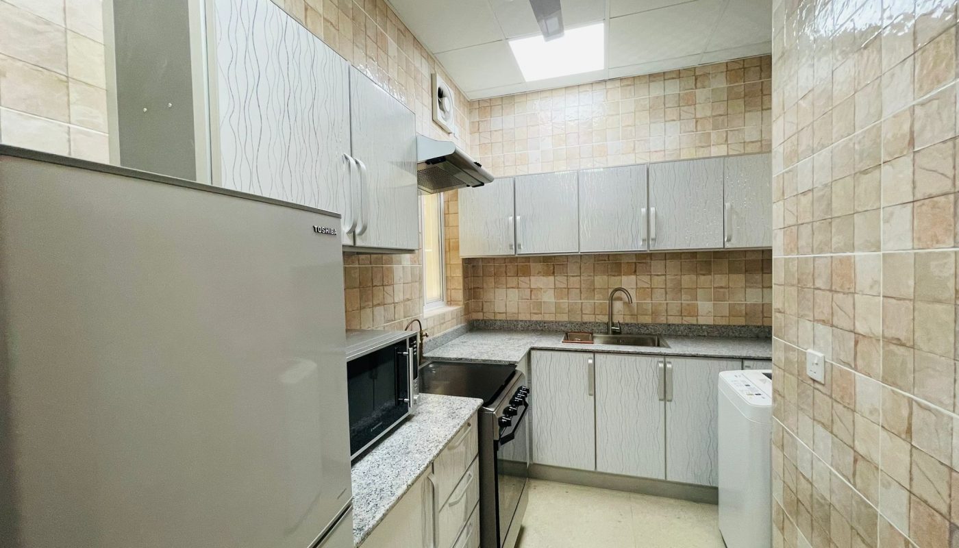 A small modern kitchen in Umm Al-Hassam, featuring beige tiled walls, grey countertops, stainless steel appliances, white cabinets, a refrigerator, oven, microwave, sink, and washing machine. This 2BR unit exudes luxury with its sleek design and high-end finishes.