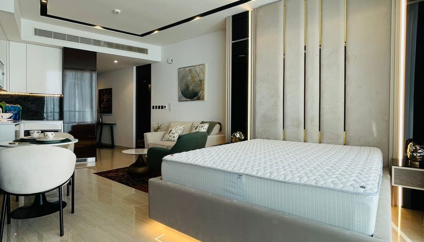 Modern bedroom with a large bed featuring under-bed lighting, a small round table with chairs, a plush rug, and a kitchenette in the background. The decor is minimalist and contemporary.