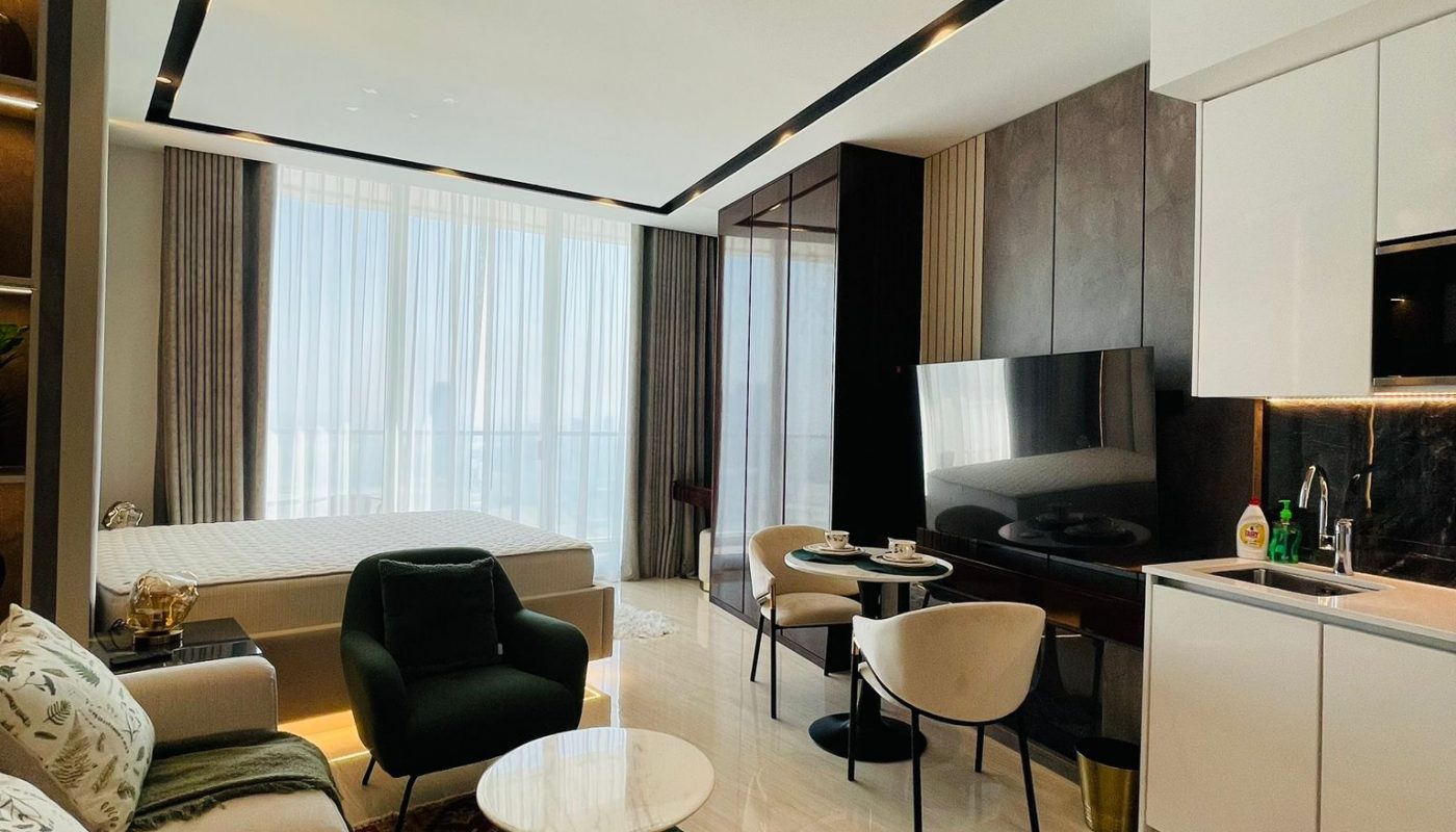 Modern studio apartment with a bed, sofa, armchairs, dining table, kitchen area, and large windows with sheer curtains, featuring neutral tones and decorative rugs.